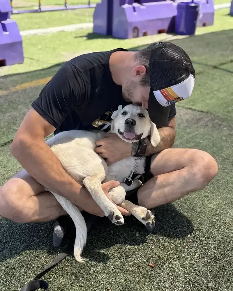 Joe sitting on the training-yard turf, hugging a white lab puppy
