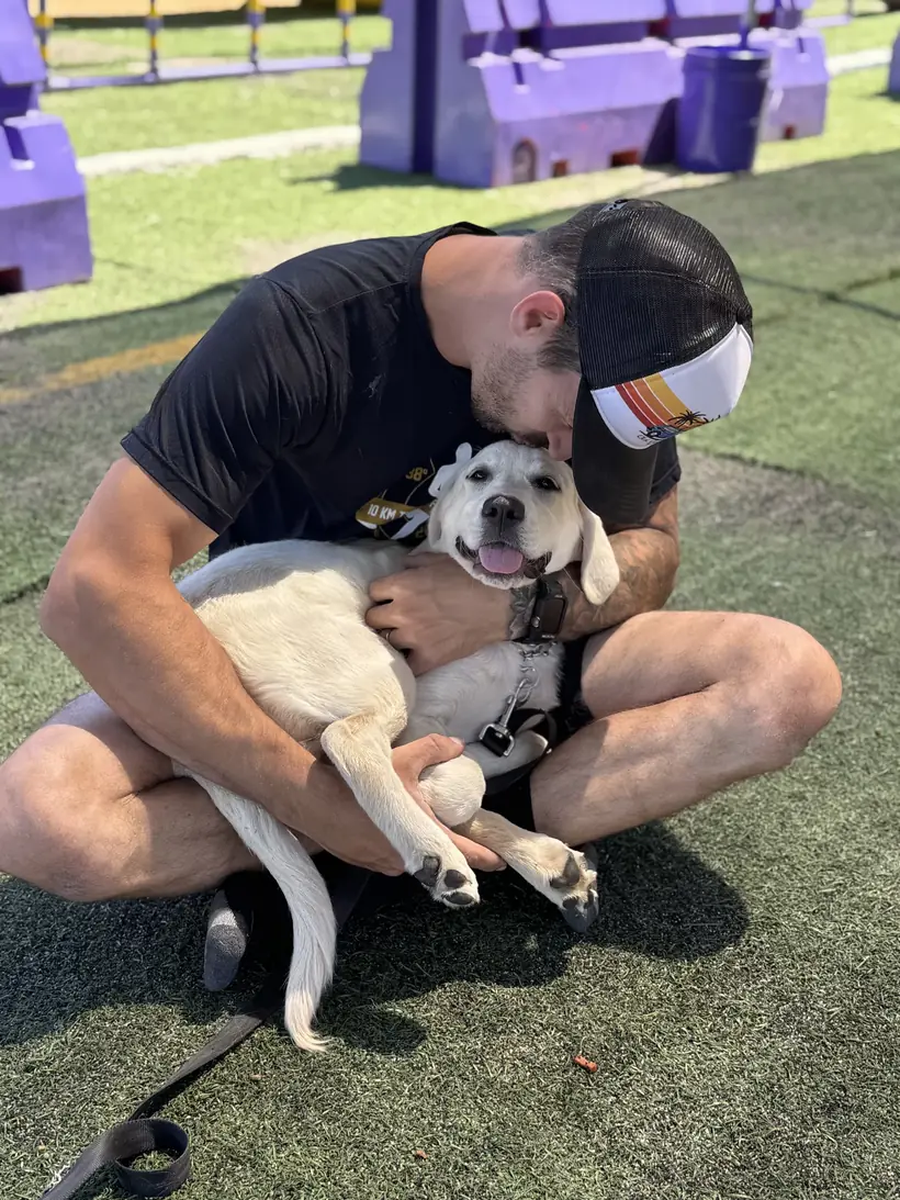 A trainer sitting on the turf hugging a white puppy