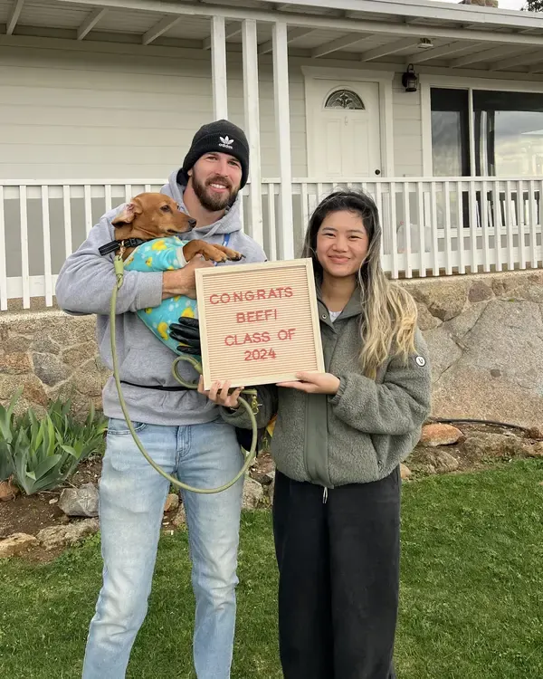 Joe holding Beef, a small red dog in duck-print pajamas, beside Beef's owner and her letter board: Congrats Beef!